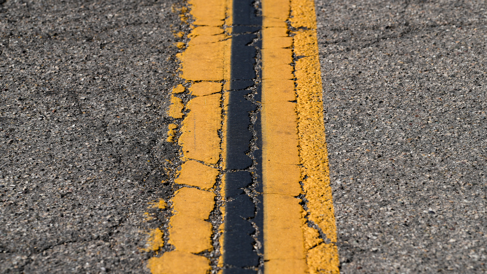 Close-up of worn double yellow centerlines on an asphalt roadway, with cracked paint and pavement deterioration visible